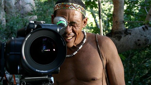 Man with a beaded headband laughing as he looks through the viewfinder of a film camera