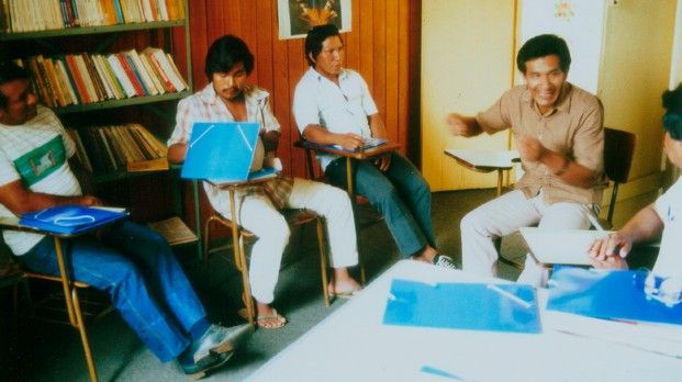Groupe d’hommes assis dans une pièce avec une bibliothèque, des classeurs bleus sur la table