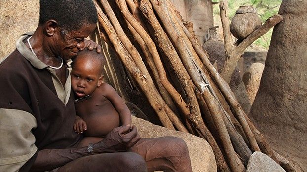 A father holding his little son in front of a mud house with wooden poles