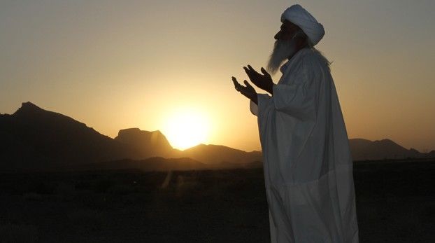 Homme en prière, vêtu de blanc et coiffé d’un turban, devant le soleil couchant sur les montagnes