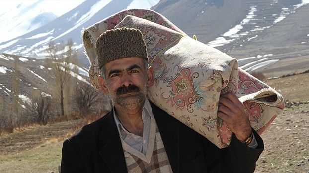 Man in a fur hat carrying a rolled-up carpet on his shoulder, mountains behind