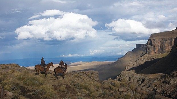 Two riders on a high plateau in front of cliffs and a cloudy sky