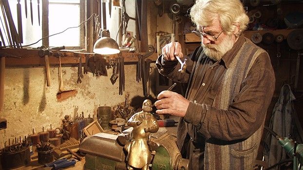 Older bearded man working on a bronze figure in a workshop full of tools
