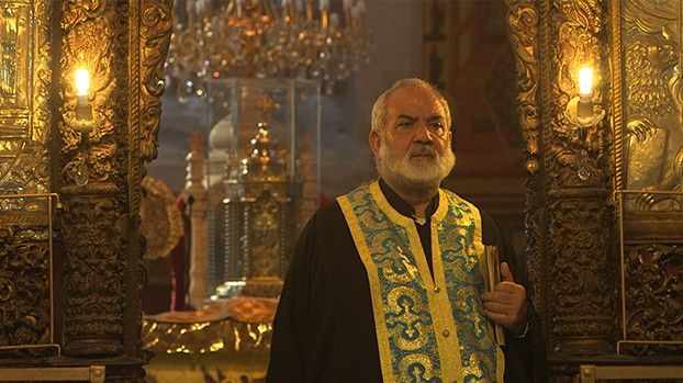 Greek Orthodox priest in gold-embroidered vestments in front of a gilded iconostasis